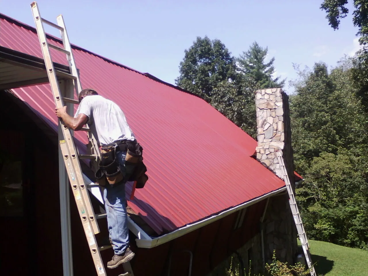 Roofer on a ladder beside a red metal roof and stone chimney — Soffit & Fascia Repair by Roofing Brothers in Leechburg